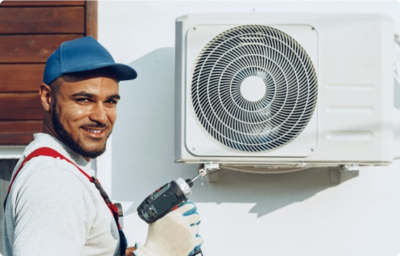 RHMH A technician from an air conditioning company in Timmins, Ontario, wearing a blue cap and gloves, uses a drill to install or repair an outdoor air conditioning unit mounted on a wall. Heating And Air Conditioning