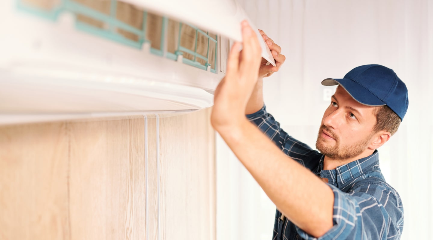 RHMH A man in a blue cap and plaid shirt installs or adjusts an air conditioning unit on a wall indoors, showcasing the expertise you'd expect from an Air Conditioning Company in Timmins Ontario. Heating And Air Conditioning