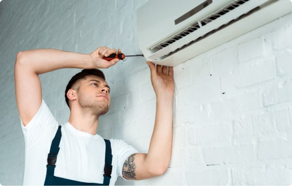 RHMH A man in a white shirt and overalls uses a screwdriver to repair or install a wall-mounted air conditioning unit for an Air Conditioning Company in Timmins, Ontario. Heating And Air Conditioning