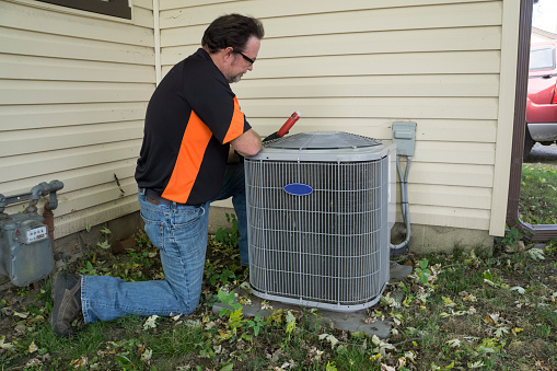 RHMH A technician in a black and orange shirt kneels beside an HVAC unit outside a house in Timmins, Ontario, representing a reputable heating company, using a tool to work on it. Heating And Air Conditioning