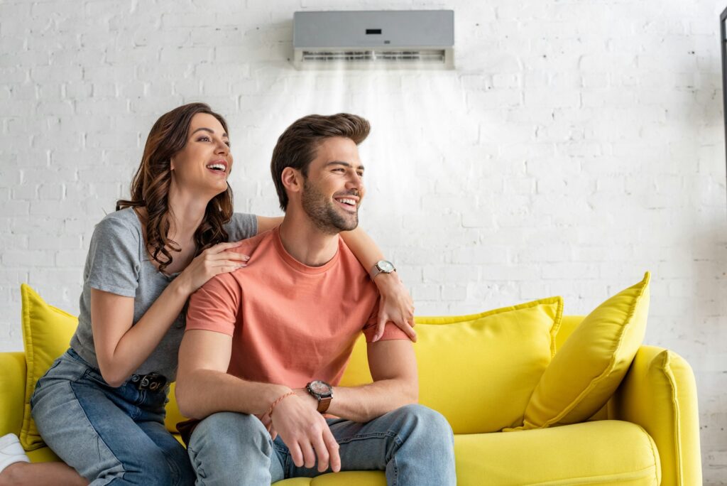 RHMH A couple sits on a yellow couch, smiling and looking to the side. A wall-mounted air conditioner above them, installed by a reputable Air Conditioning Company in Timmins, Ontario, is running efficiently. Heating And Air Conditioning