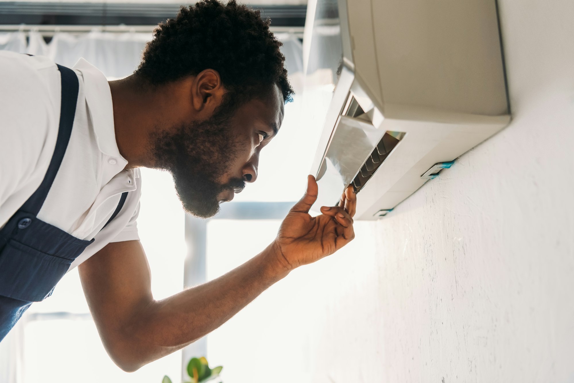 RHMH A man in a white polo shirt and blue overalls inspects an air conditioning unit mounted on a wall, showcasing the expertise of our Air Conditioning Company in Timmins, Ontario. Heating And Air Conditioning