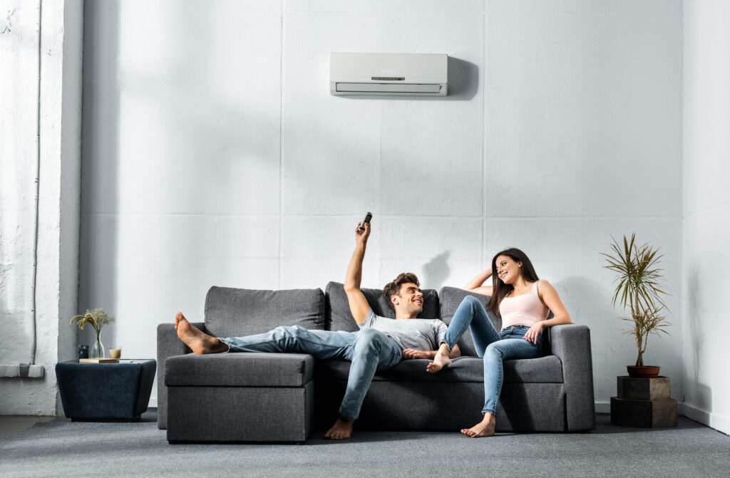 RHMH A man and a woman relax on a gray sofa, with the man holding a remote control and looking at the woman. An air conditioner from an Air Conditioning Company in Timmins, Ontario is mounted on the wall above them. Heating And Air Conditioning