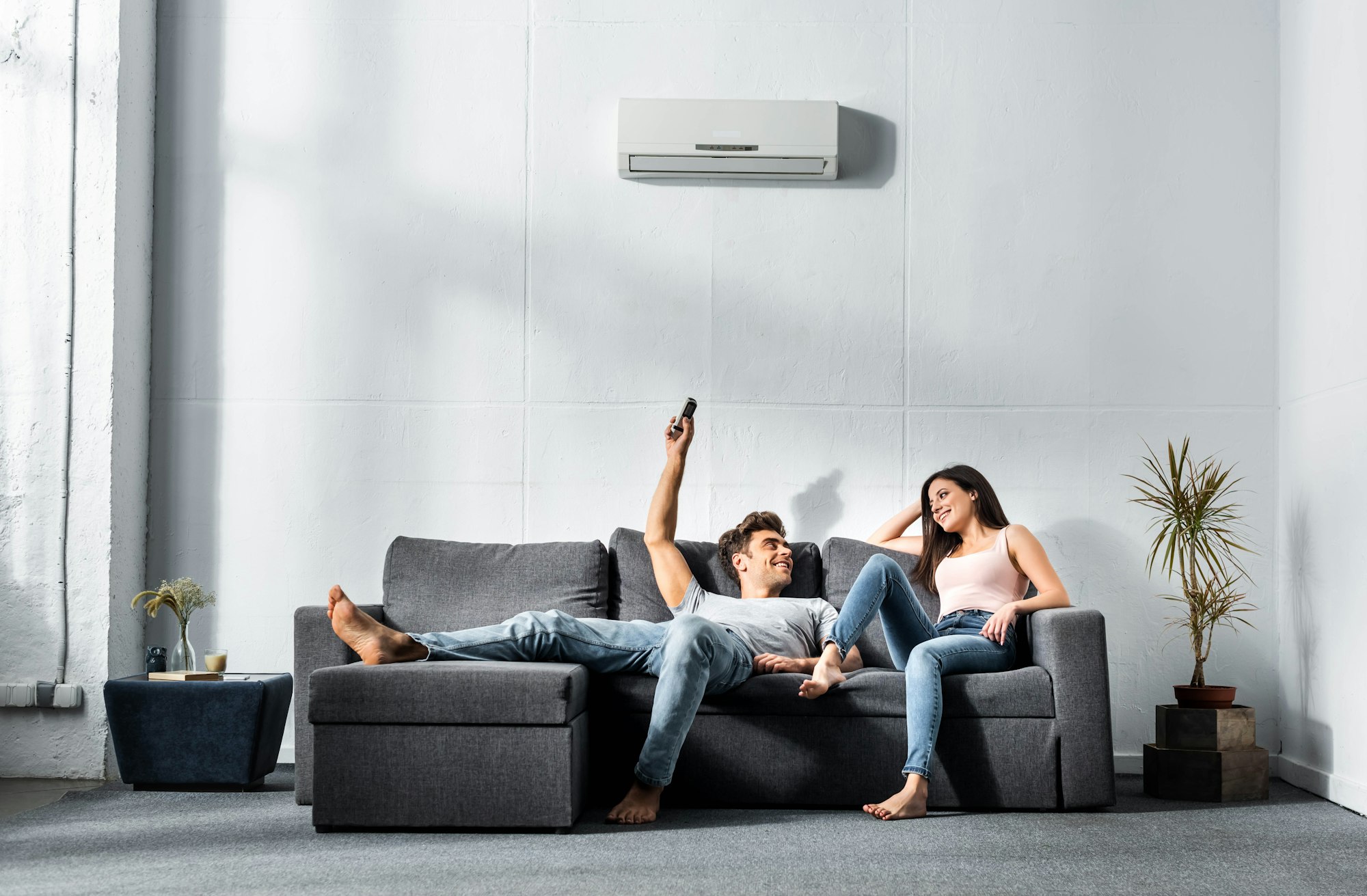 RHMH A man and a woman relax on a gray sofa, with the man holding a remote control and looking at the woman. An air conditioner from an Air Conditioning Company in Timmins, Ontario is mounted on the wall above them. Heating And Air Conditioning