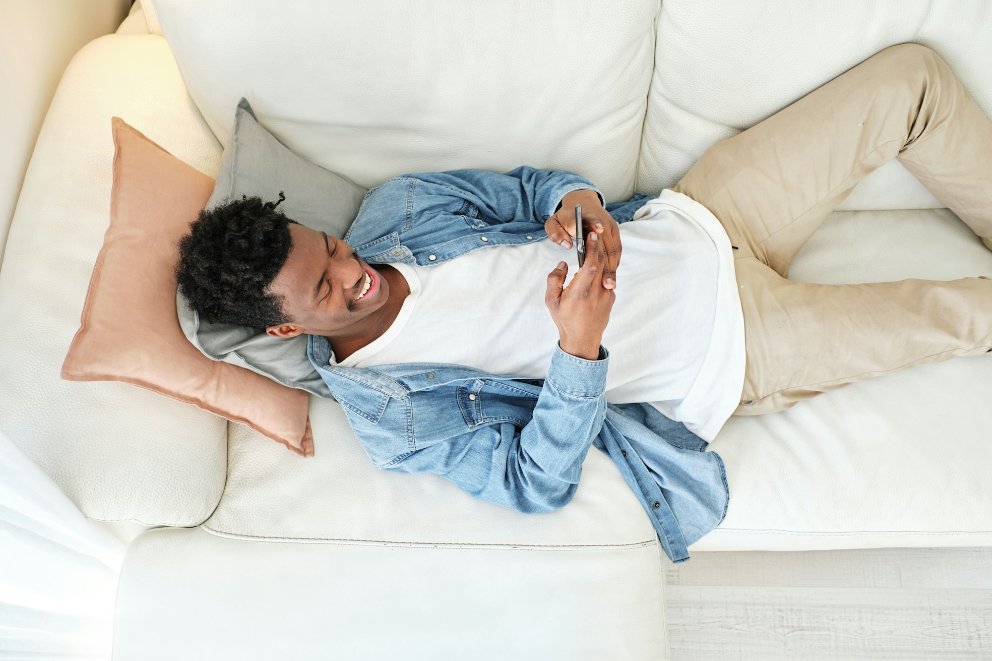 RHMH A young man is lying on a white couch, using a smartphone and smiling. Dressed in beige pants, a white t-shirt, and a denim shirt, he relaxes against light-colored pillows. Perhaps he's browsing for the best heating company in Timmins, Ontario. Heating And Air Conditioning