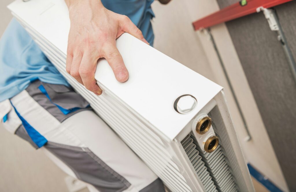 RHMH A person in blue and gray work clothes, likely from a heating company in Timmins Ontario, is installing a white radiator with two brass connection ports and a silver knob. Heating And Air Conditioning