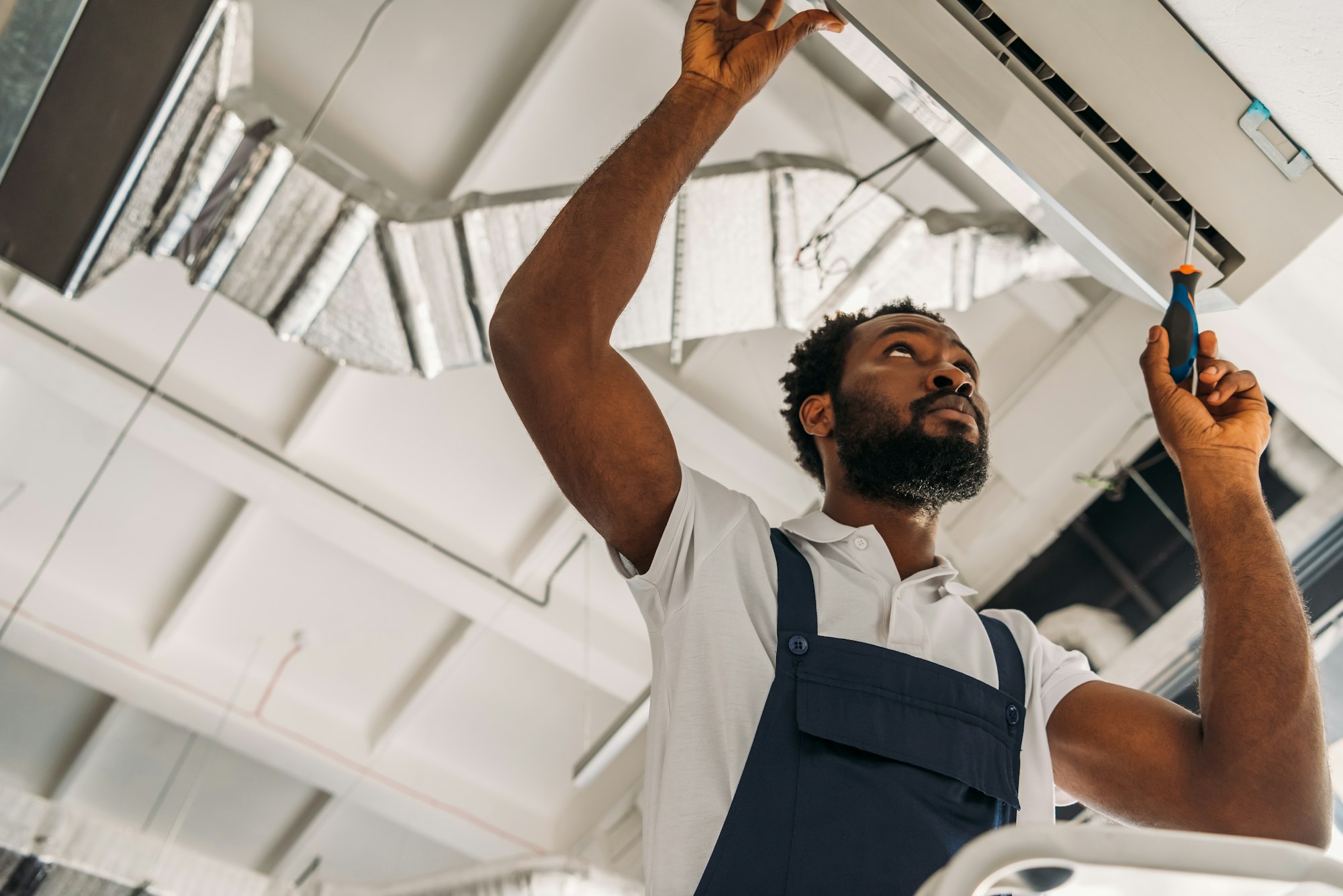 RHMH A technician in blue overalls and a white shirt, representing an Air Conditioning Company in Timmins, Ontario, uses a screwdriver to repair an overhead air conditioning unit. Heating And Air Conditioning