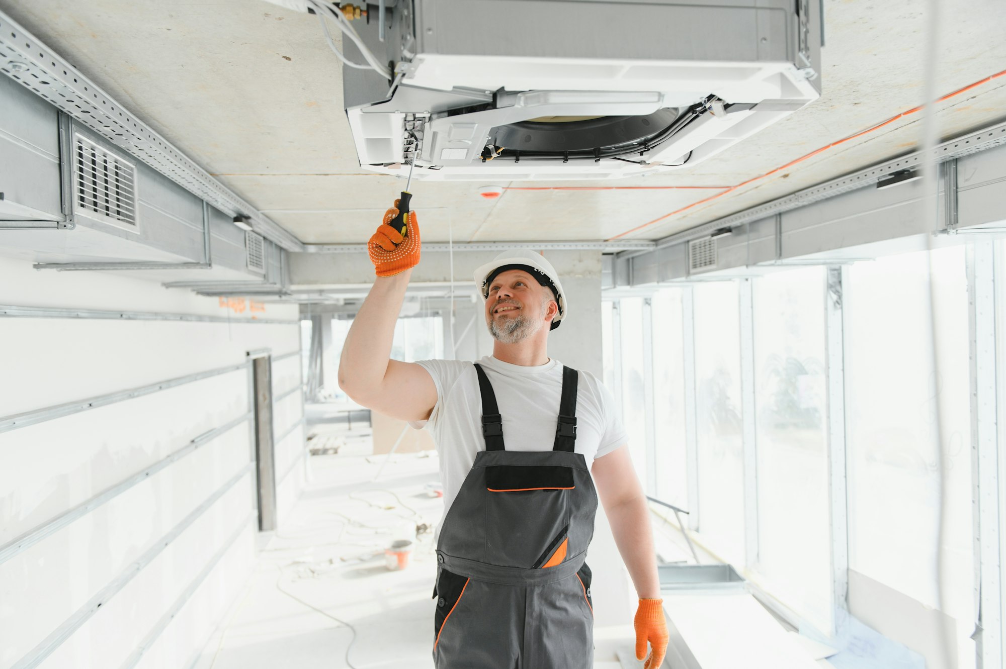 RHMH A construction worker in overalls and a white helmet adjusts a ceiling-mounted air conditioning unit in a well-lit, unfinished building interior for an Air Conditioning Company based in Timmins, Ontario. Heating And Air Conditioning