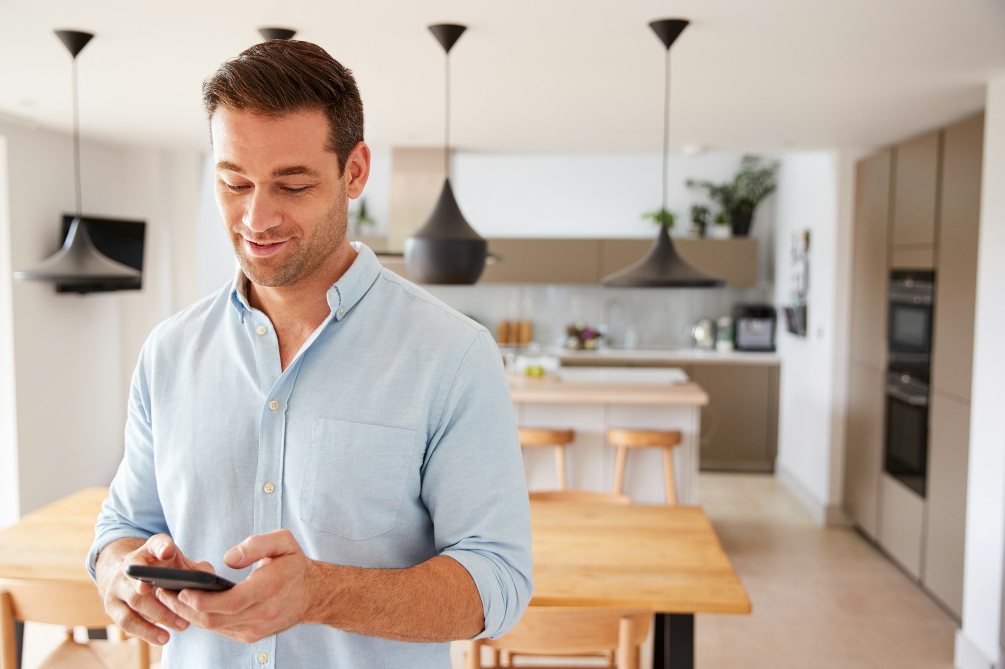 RHMH A man stands in a modern kitchen in Timmins, Ontario, looking at his smartphone and wearing a light blue shirt. The kitchen features a wooden dining table and contemporary lighting, perhaps checking the latest service updates from his local air conditioning company. Heating And Air Conditioning