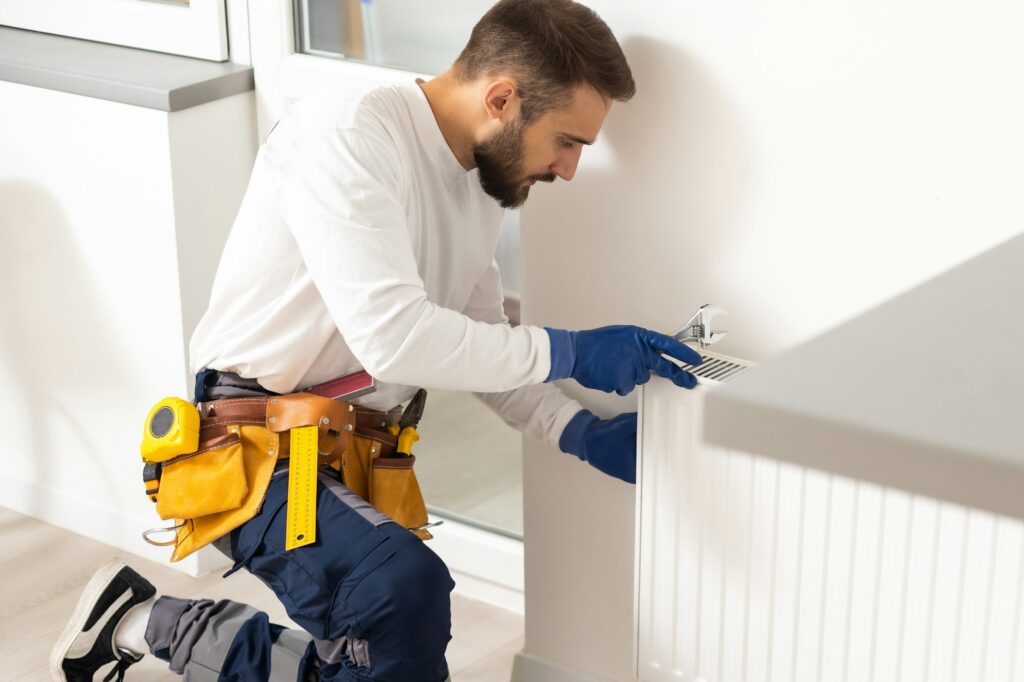 RHMH A technician from a leading Heating Company in Timmins, Ontario, kneels to adjust a wall-mounted radiator. He wears a tool belt and blue protective gloves, focusing on his task with a concentrated expression. Heating And Air Conditioning