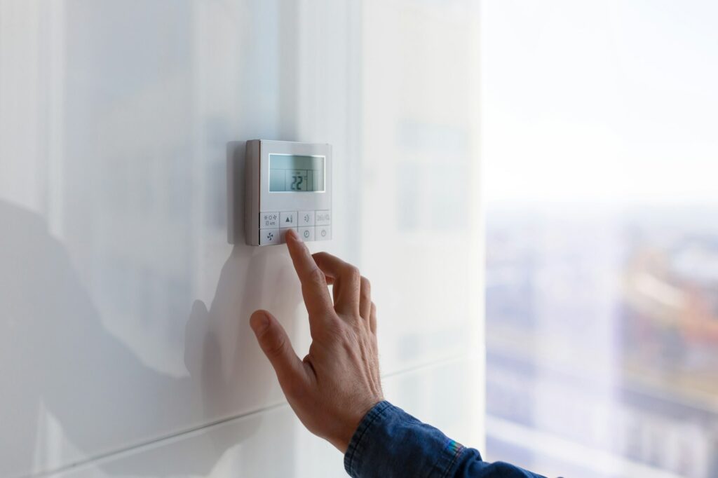 RHMH A hand adjusting a wall-mounted digital thermostat displaying 22°C in a bright room, representing the reliable services provided by an Air Conditioning Company in Timmins, Ontario. Heating And Air Conditioning
