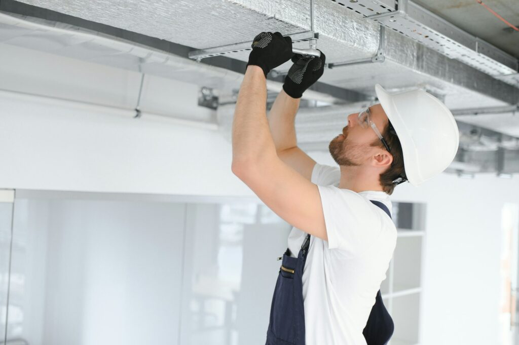 RHMH A construction worker wearing a white hard hat and black gloves uses a wrench to adjust a metal fixture on a ceiling for an air conditioning company in Timmins, Ontario. Heating And Air Conditioning
