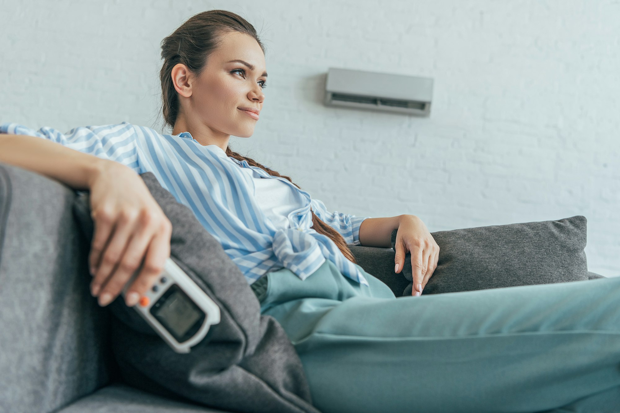 RHMH A woman lounges on a couch holding an air conditioning remote, looking off to the side, in a bright, modern living room with a white brick wall. The impeccable comfort provided by Timmins Ontario's premier Air Conditioning Company is evident in every detail of her serene surroundings. Heating And Air Conditioning